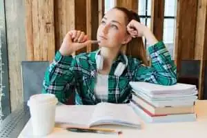 Young woman in a plaid shirt with headphones, thoughtfully contemplating while studying at a table with books and a coffee cup, reflecting the importance of effective learning environments in schools.