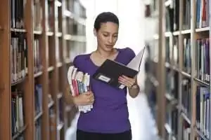Woman in a library holding books and reading a document, symbolizing knowledge acquisition and research for ISO 42001 certification preparation.
