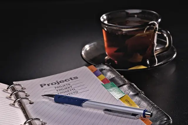 Notizbuch mit dem Titel "Projects", blauer Stift und Tasse Tee, symbolisiert Projektmanagement und Planung für ISO 27001 Zertifizierungen.